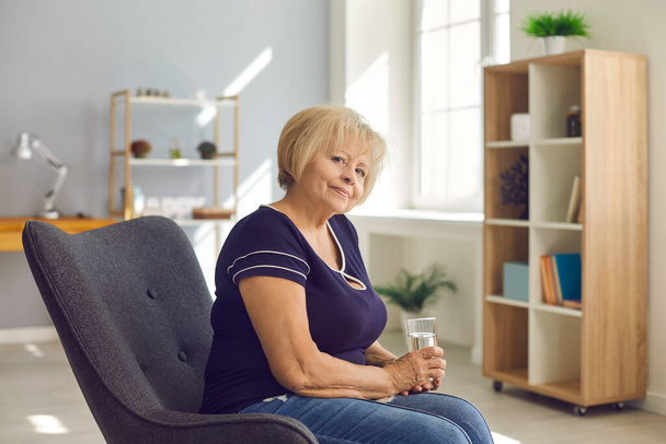 Pozytywny starszy emeryt babcia w dżinsach i t-shirt siedzi ze szklanką wody w fotelu - Zdjęcie, obraz