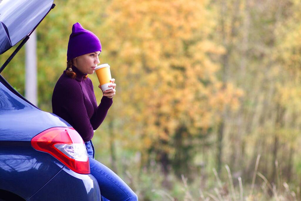 Young beautiful woman drinking coffee or tea from paper cup sitting, leaning on a car trunk in a golden autumn forest in a cold weather in hat and sweater. Traveling by car. - Foto, imagen