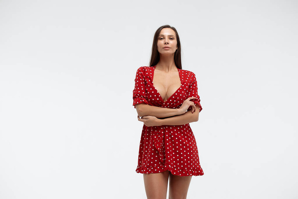 セクシー女性身に着けています赤い短いポルカドットドレス立っています上の白い背景でスタジオとタッチ毛の中にカメラを見て - 写真・画像