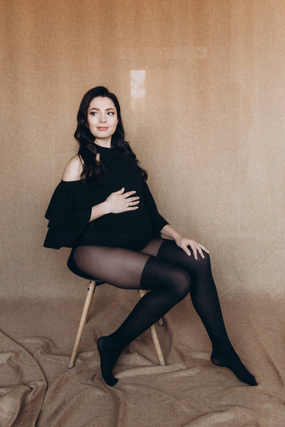 Pregnancy photo session in a photo studio with a fabric background in a black bodysuit and tights - Foto, Imagem