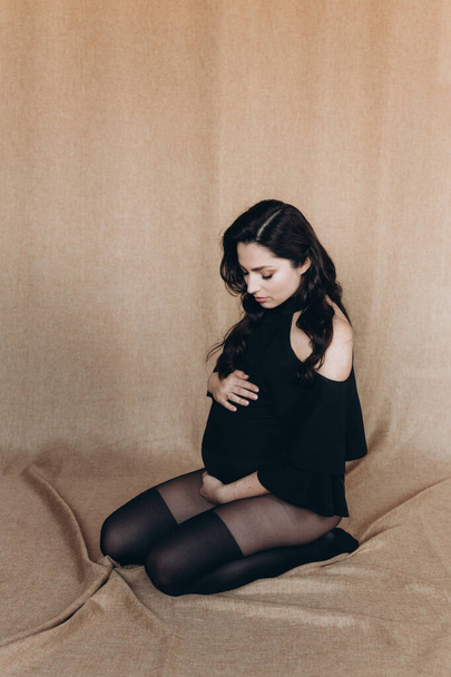 Sessione fotografica di gravidanza in uno studio fotografico con uno sfondo di tessuto in un body nero e collant - Foto, immagini