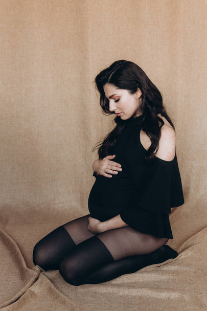Sessione fotografica di gravidanza in uno studio fotografico con uno sfondo di tessuto in un body nero e collant - Foto, immagini