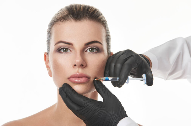 Красивая молодая женщина лицо и рука в перчатке со шприцем делает инъекцию на губах. Изолированный на белом фоне. Интимационная терапия красоты - Фото, изображение