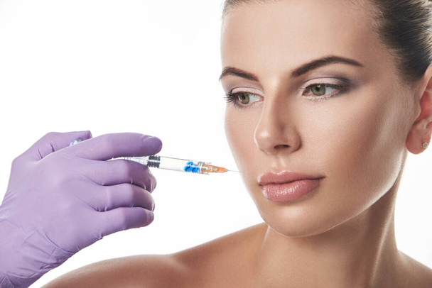 Beautician's hand in purple glove holding a syringe near beautiful woman's face. Portrait isolated on a white background. - Foto, Imagem