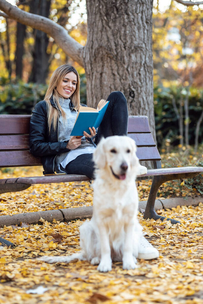 Shot of beautiful young woman reading a book while sitting on bench with her lovely golden retriever dog in the park in autumn. - Photo, image