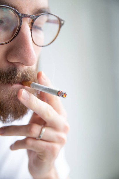 Nicotine addiction. Young adult, bearded man in a white t-shirt smoking a cigarette, close-up. Quarantine at home.Depression. Smoking break during a working day.  - Foto, afbeelding