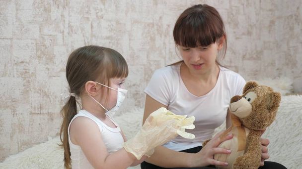 Ein Kind in medizinischer Maske zieht Gummihandschuhe an und spielt mit seiner Schwester. Kinder behandeln einen Spielzeugbär. Das Kind spielt im Krankenhaus. Traum, Arzt, Krankenpfleger, Tierarzt zu werden und Kranke zu heilen. - Foto, Bild