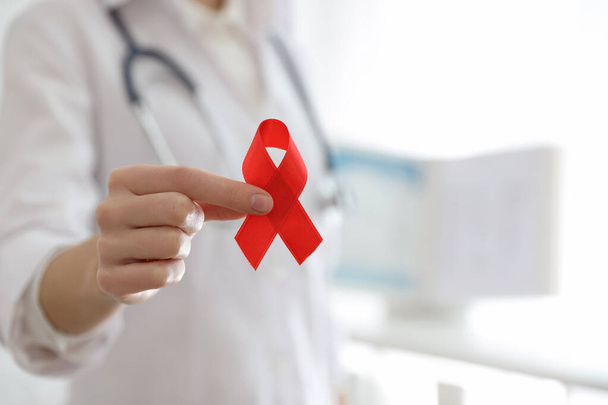 Doctor holding red ribbon on blurred background, closeup with space for text. AIDS awareness month - Foto, Bild