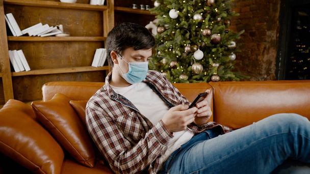 Молодий сумний чоловік в медичній масці читає новини зі смартфона вдома на дивані біля ялинки в карантині через коронавірус. Погані новини, хвороба. Святкування на самоті. Замкнення ковадла 19
. - Фото, зображення