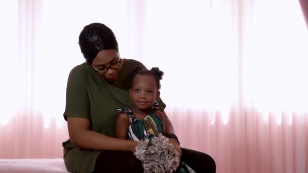 Afrikanisches Mädchen hält eine Blume in der Hand, wenn die Mutter ihre Haare setzt. Glückliche Familie Afrikanische Mutter und Kind spielen zusammen, während sie im Schlafzimmer auf dem Bett sitzen. - Filmmaterial, Video