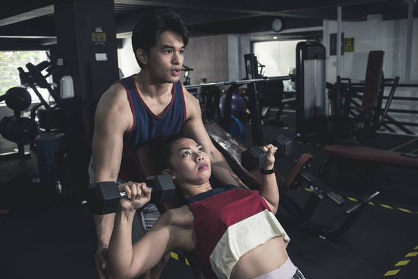 Un joven asiático que ve a una chica atlética haciendo prensas de mancuernas inclinadas. Ejercicio con asistencia en el gimnasio. - Foto, Imagen