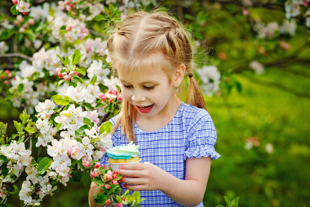 Egy gyermek egy virágzó almás gyümölcsösben élvezi a meleg tavaszi napot. A lány egy tortát tart a kezében, meg akarja enni.. - Fotó, kép