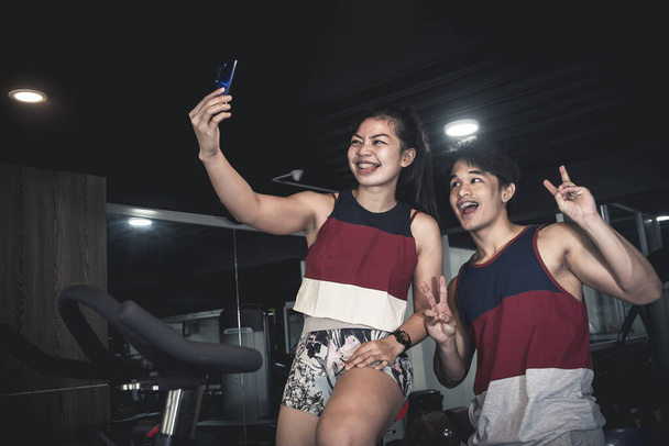 Dos amigos asiáticos o una pareja con camisetas de tanque a juego se toman unos selfies en el gimnasio. La chica está sentada en una bicicleta estacionaria. Entorno interior atenuado - Foto, imagen