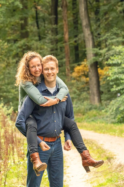 Attractive young couple, man carries woman on his back in the woods. They stand still on a sandy forest path - 写真・画像