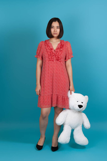 full-length portrait of a sad, upset, offended, lonely woman in a red dress holding a white Teddy bear by the ear, isolated on a blue background - Photo, Image
