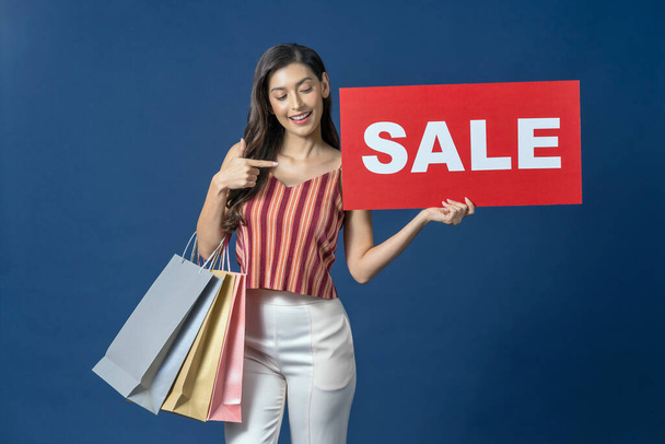 Happy young Asian woman holding sales banner and pointing to sales paper banner for shopping on blue color background, cashless payment, black friday and consumerism concept, carrying shopping bags - Photo, Image