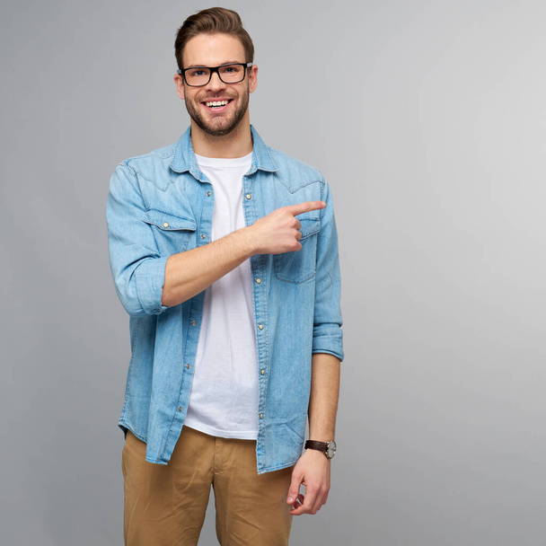 Happy young handsome man in jeans shirt pointing away standing against grey background - Photo, image