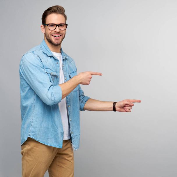 Happy young handsome man in jeans shirt pointing away standing against grey background - Фото, зображення