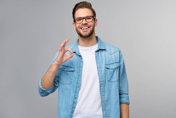 Portrait of young handsome caucasian man in jeans shirt showing ok sign gesture standing over light background - 写真・画像
