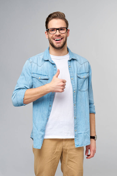 Portrait of young handsome caucasian man in jeans shirt showing big thumb up gesture standing over light background - Zdjęcie, obraz