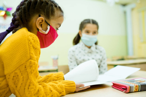 Meisjes met beschermende maskers zitten aan hun bureau in de les en lezen het lesboek - Foto, afbeelding