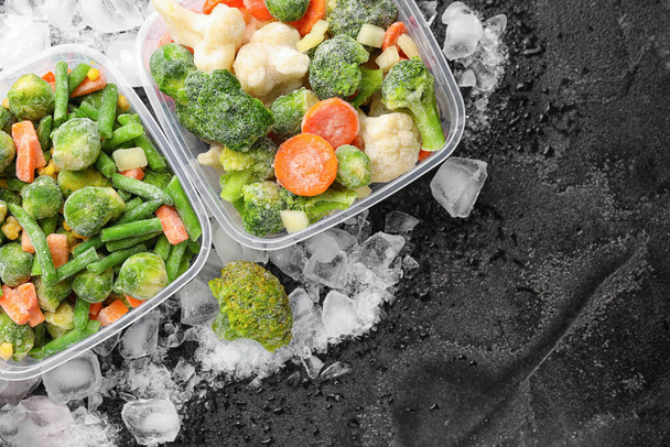 Envases de plástico con verduras congeladas y hielo sobre fondo oscuro - Foto, Imagen