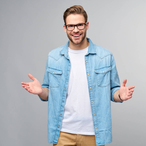 young man wearing jeans shirt welcoming you with a smile on his face and his arms wide open standing over grey background - 写真・画像