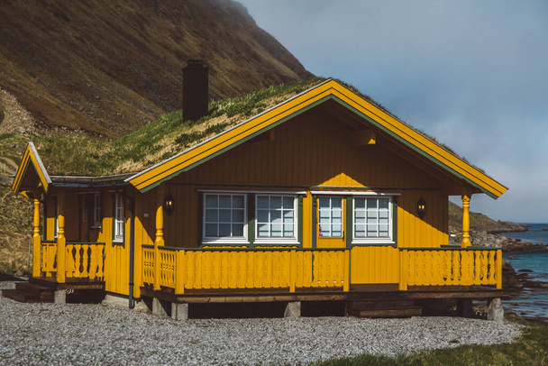 Geel houten huis met mos op het dak tegen de achtergrond van de zee en de bergen - Foto, afbeelding