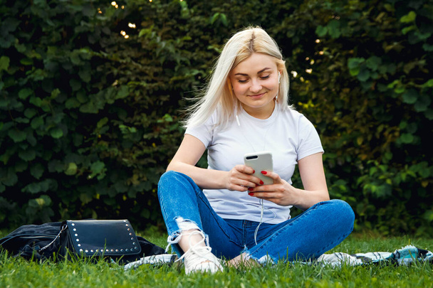 ヘッドフォンや携帯電話の美しいブロンドの女の子は緑の芝生の上に座っています。彼女はヘッドフォンで電話で話している。ライフスタイルの概念。テキストのコピー、空のスペース. - 写真・画像
