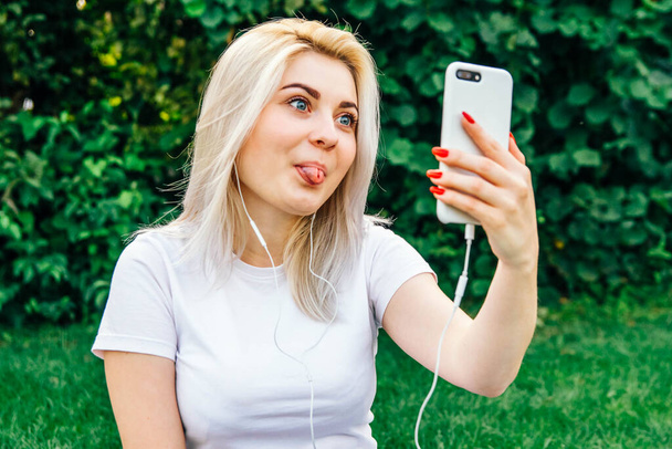Portret van mooi blond meisje in hoofdtelefoon en smartphone in handen zittend op groene achtergrond. Ze praat aan de telefoon via een koptelefoon. Tijd om selfie te nemen. Kopieer, lege ruimte voor tekst. - Foto, afbeelding