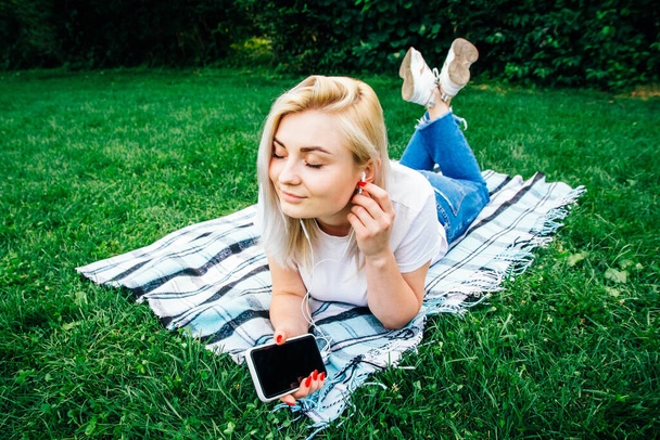 Krásná blondýnka ve sluchátkách a smartphone v ruce poslouchá hudbu ležící na zeleném pozadí. Koncept životního stylu. Kopírovat, prázdné místo pro text. - Fotografie, Obrázek