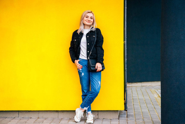Schöne blonde Mädchen in einer schwarzen Jacke posiert vor einem Hintergrund der gelben Wand. Kopieren, leerer Raum für Text. - Foto, Bild