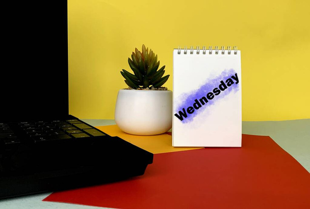Τετάρτη σε ένα λευκό σημειωματάριο, δίπλα σε ένα γλάστρα λουλούδι, φορητό υπολογιστή σε γραφείο γραφείου.Ημερολόγιο για κάθε μέρα της εβδομάδας. - Φωτογραφία, εικόνα