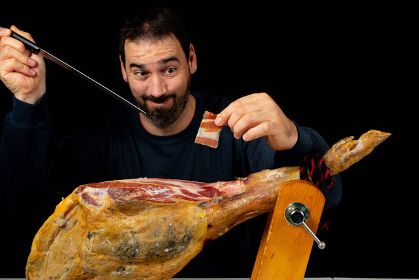 Bearded man with a ham knife ready to cut a piece of typical Spanish ham isolated on black studio background. Super foods. Delicious appetizer. Feeding concept - Photo, Image