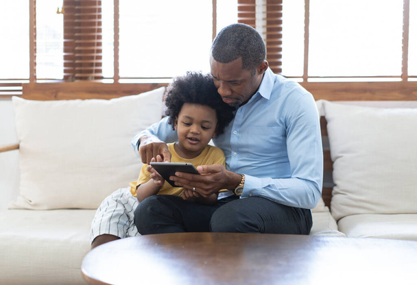幸せなアフリカ系アメリカ人の父親と息子の家でソファに座っている間、タブレットを使用しての肖像画。一緒に素晴らしい時間を過ごす。家族の娯楽、教育、技術の概念. - 写真・画像