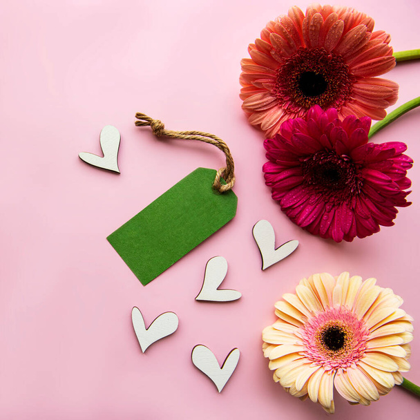 Gerbera çiçekleri, tahta kalpler ve pembe pastel arka planda yeşil kağıt etiket. Üst görünüm - Fotoğraf, Görsel