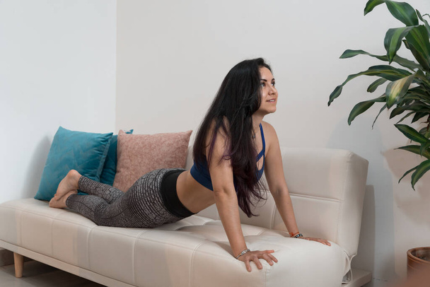 belle jeune femme faisant des exercices d'étirement sur un canapé blanc avec des coussins colorés à l'intérieur de sa maison avec une plante feuillue, yoga - Photo, image