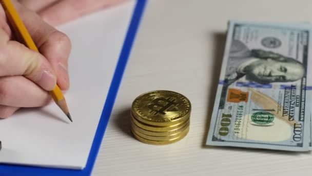 Ein Händler für digitale Währungen schreibt die Werte des Bitcoin-Kurses auf Papier nieder, es gibt Bitcoin-Münzen und Dollarscheine in der Nähe. - Filmmaterial, Video