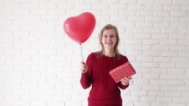 Νεαρή ξανθιά γυναίκα με κόκκινο πουλόβερ κρατώντας ένα κουτί δώρου και μπαλόνι κόκκινη καρδιά γιορτάζει την ημέρα του Αγίου Βαλεντίνου - Πλάνα, βίντεο