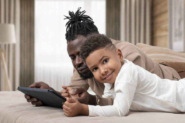 Hombre africano de piel oscura y su pequeño hijo pasan tiempo juntos. Se tumban en el sofá en casa y usan tabletas. Sonriente chico mirando a la cámara. - Foto, Imagen