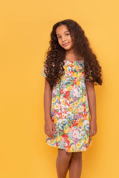 Половина портрета темнокожей молодой девушки с длинными вьющимися волосами в красочном платье на жёлтом фоне - Фото, изображение
