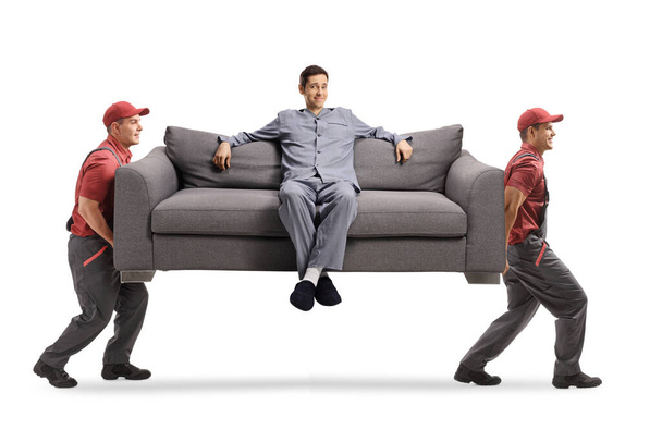 Movers carrying a sofa with a young man in pajamas sitting  isolated on white background - Photo, Image