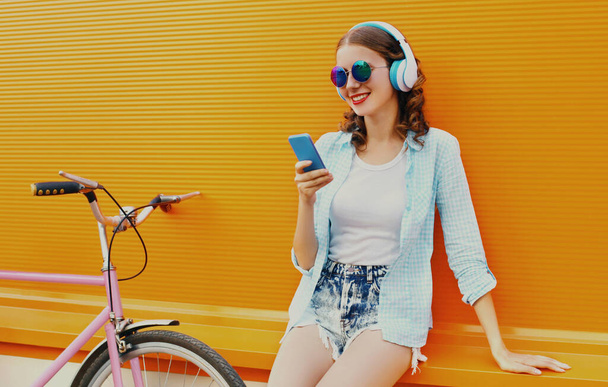 Portrait of happy young smiling woman with phone listening to music in headphones with bicycle on an orange background - Fotó, kép