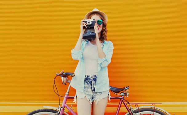 Happy young woman with film camera and bicycle on an orange background - Foto, Imagen