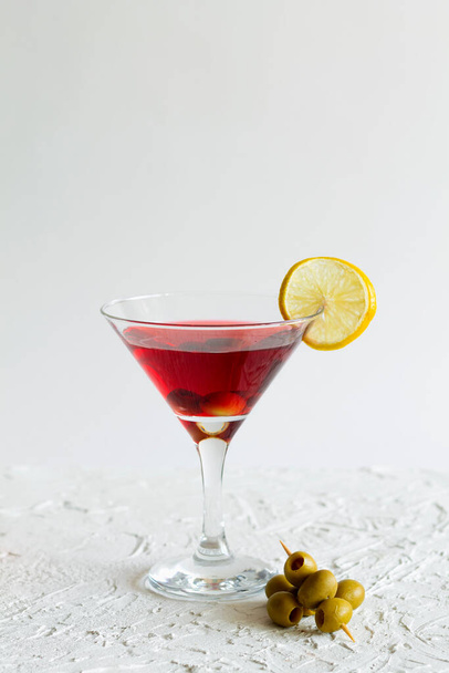 kosmopolitní koktejl v baru. Martini sklenice červeného koktejlu s olivami a citrónem na bílém pozadí. Brýle na martini na stole. kopírovat prostor. alkoholické nápoje. vertikální - Fotografie, Obrázek
