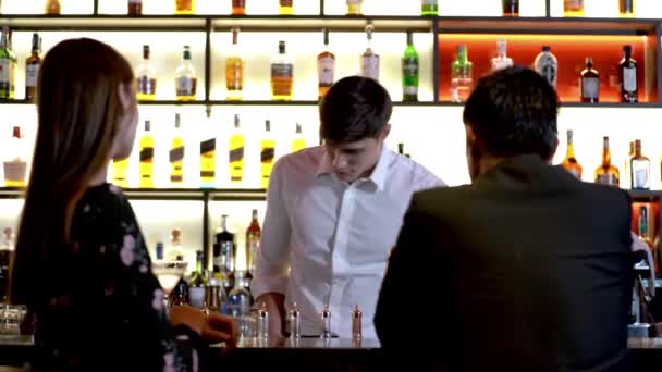 Young man smiling bartender professionally pour drink to shaker using beaker. couple of people sitting at counter bar to drink for celebrate. Different bottles and glasses stand on bar counter. - Footage, Video