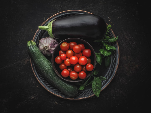 Eggplant, zucchini, garlic, cherry tomatoes and basil on vintage metal plate. Overhead shot with copy space. Dark and moody. - Foto, afbeelding