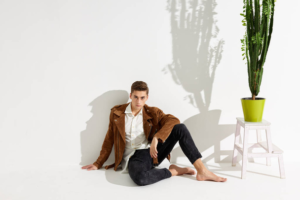 homem bonito na jaqueta marrom e calças senta-se no chão perto de uma flor em um pote - Foto, Imagem