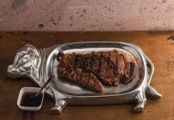 Cut of meat "Aguja nortea Sonora", served on a silver plate in the shape of a cow, the meat is in half, covered by chimichurri, on a wooden background and quarry - Photo, Image