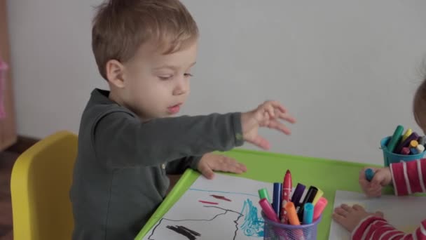 Sanat, eğitim, çocukluk, kavramlar- küçük mutlu okul öncesi küçük çocuk keçeli kalem ve kalemlerle çizim yapıyor kapalı bir masada oturuyor. Güler yüzlü çocuklar, kardeşim içeride resim yapar. - Video, Çekim